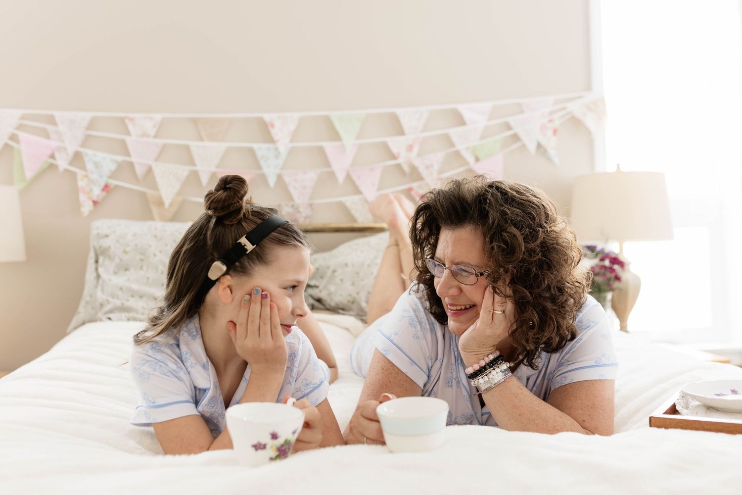 Mother and daughter in pajamas lying on bed sharing morning tea, smiling and talking in cozy bedroom.
