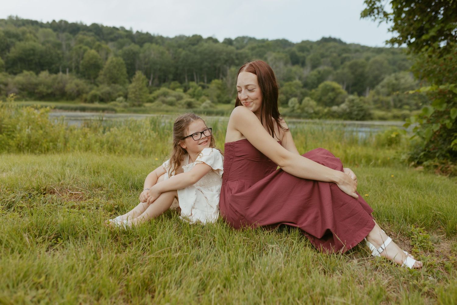 A mother and daughter sit together on green grass near a lake, surrounded by lush trees on a cloudy day.