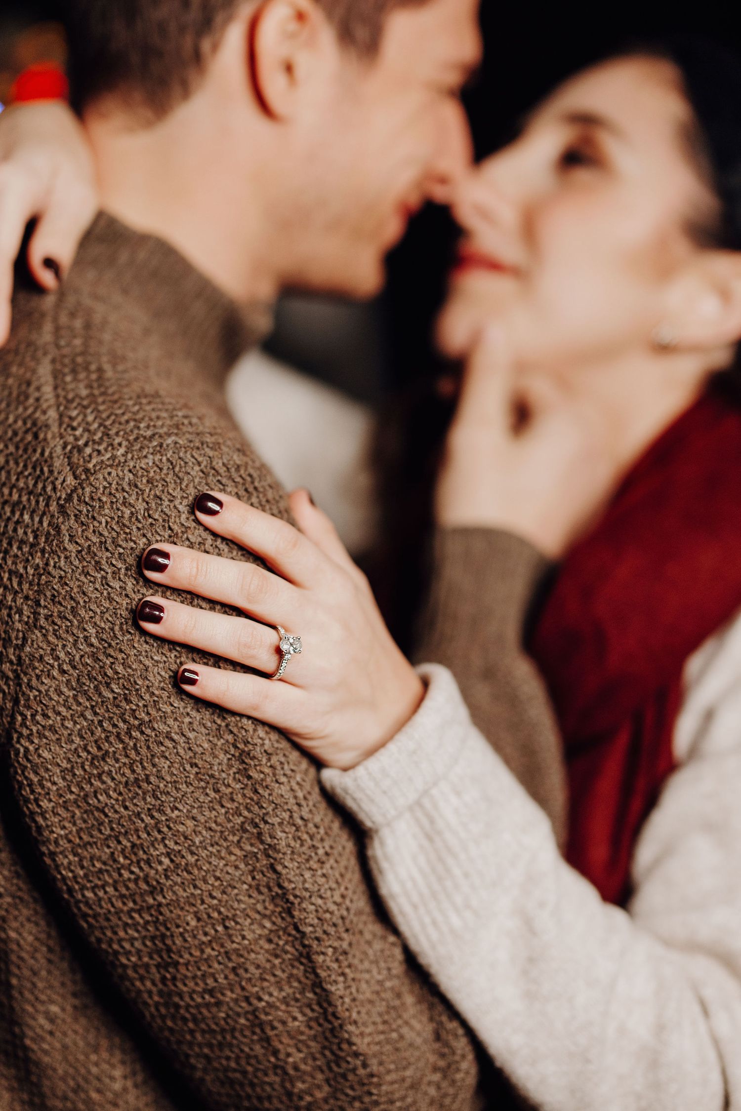 Close-up of a woman's hand with dark nails wearing a diamond engagement ring resting on a man's brown sweater.