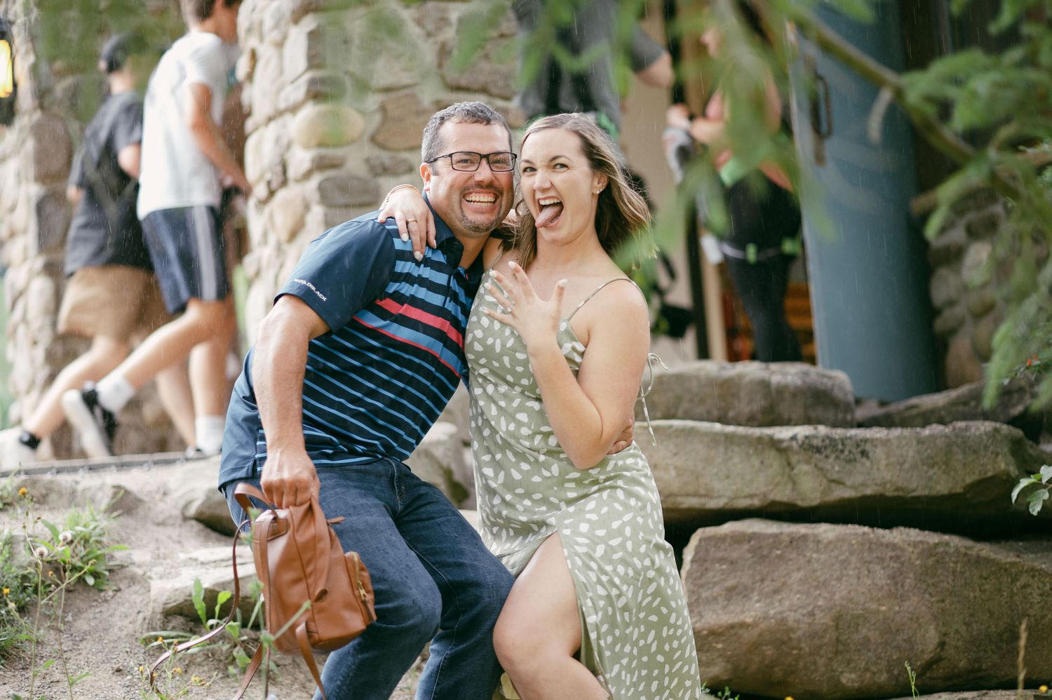 A laughing couple sits on rocks outdoors, man in striped shirt with bag, woman in floral dress, tropical setting.