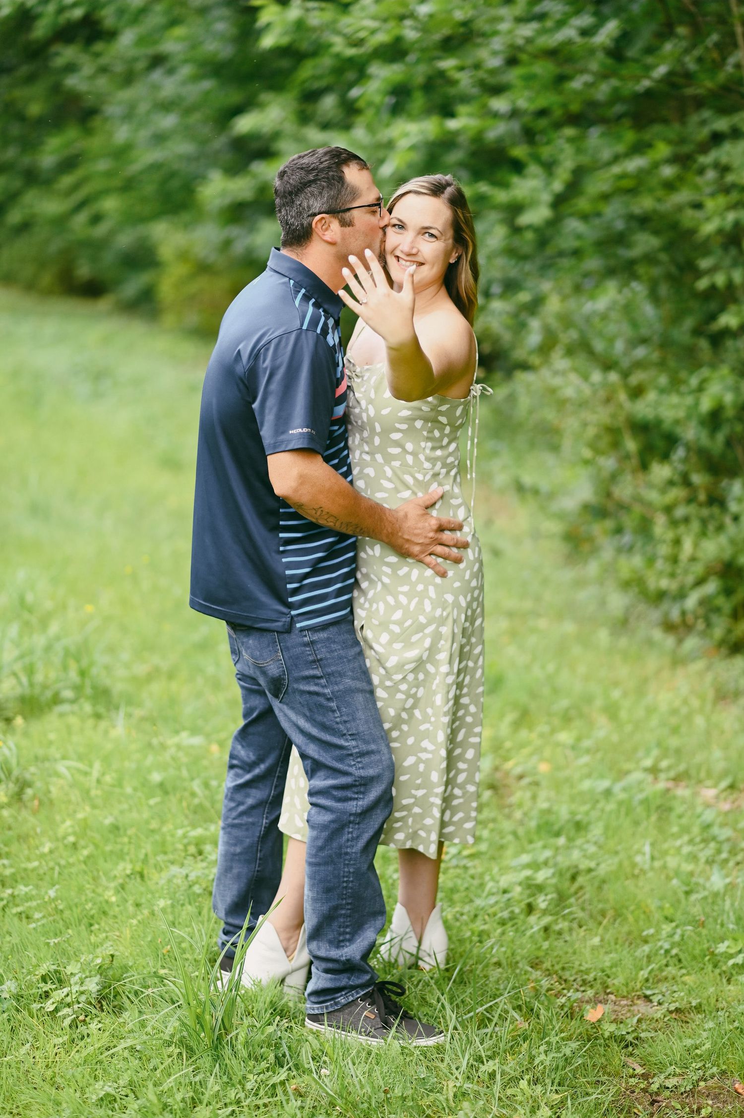 Couple embracing outdoors, woman showing engagement ring while smiling, surrounded by lush green trees.