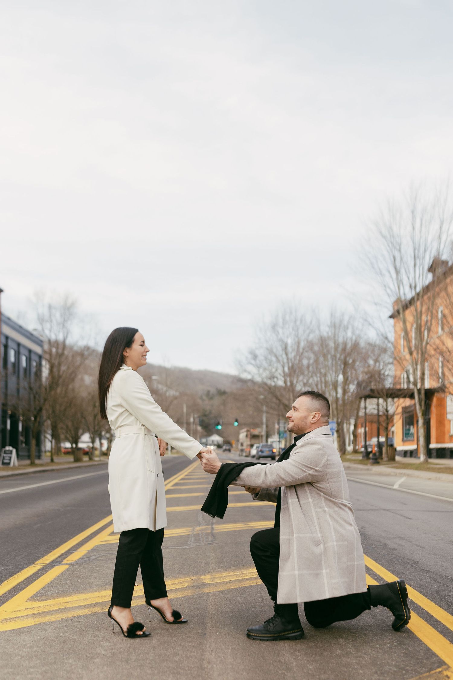 Man proposes on one knee to woman on a city street, both wearing elegant coats on a cloudy day.