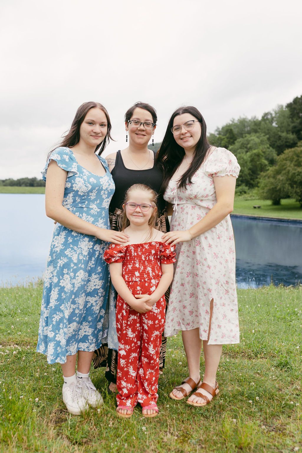Three people in floral dresses pose together on a sunny grassy field near a lake.