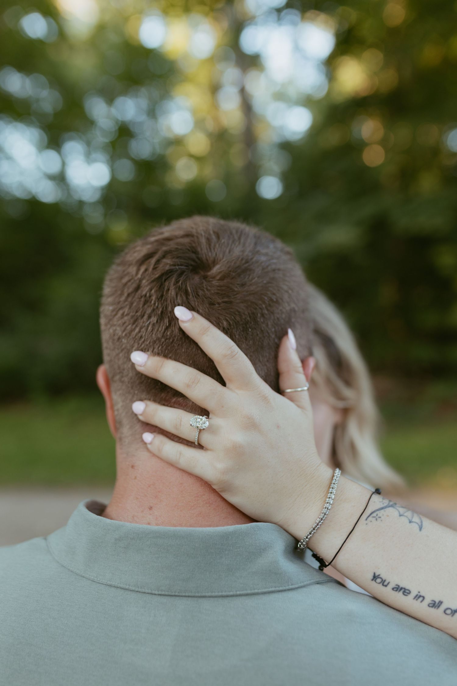 A woman's hand with an engagement ring and tattoo rests on a man's neck outdoors.