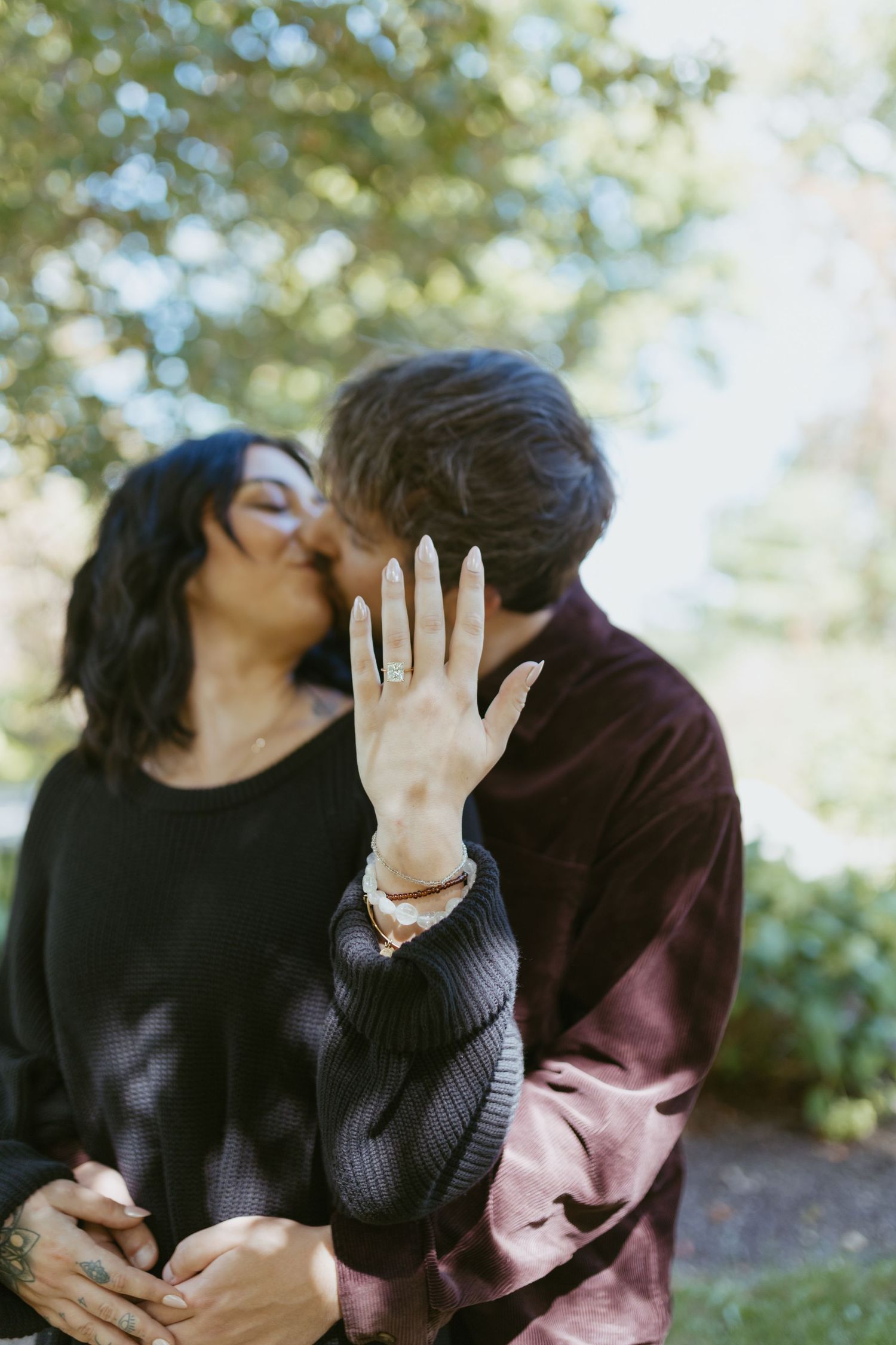 Couple sharing a kiss while showing off an engagement ring outdoors near green trees.