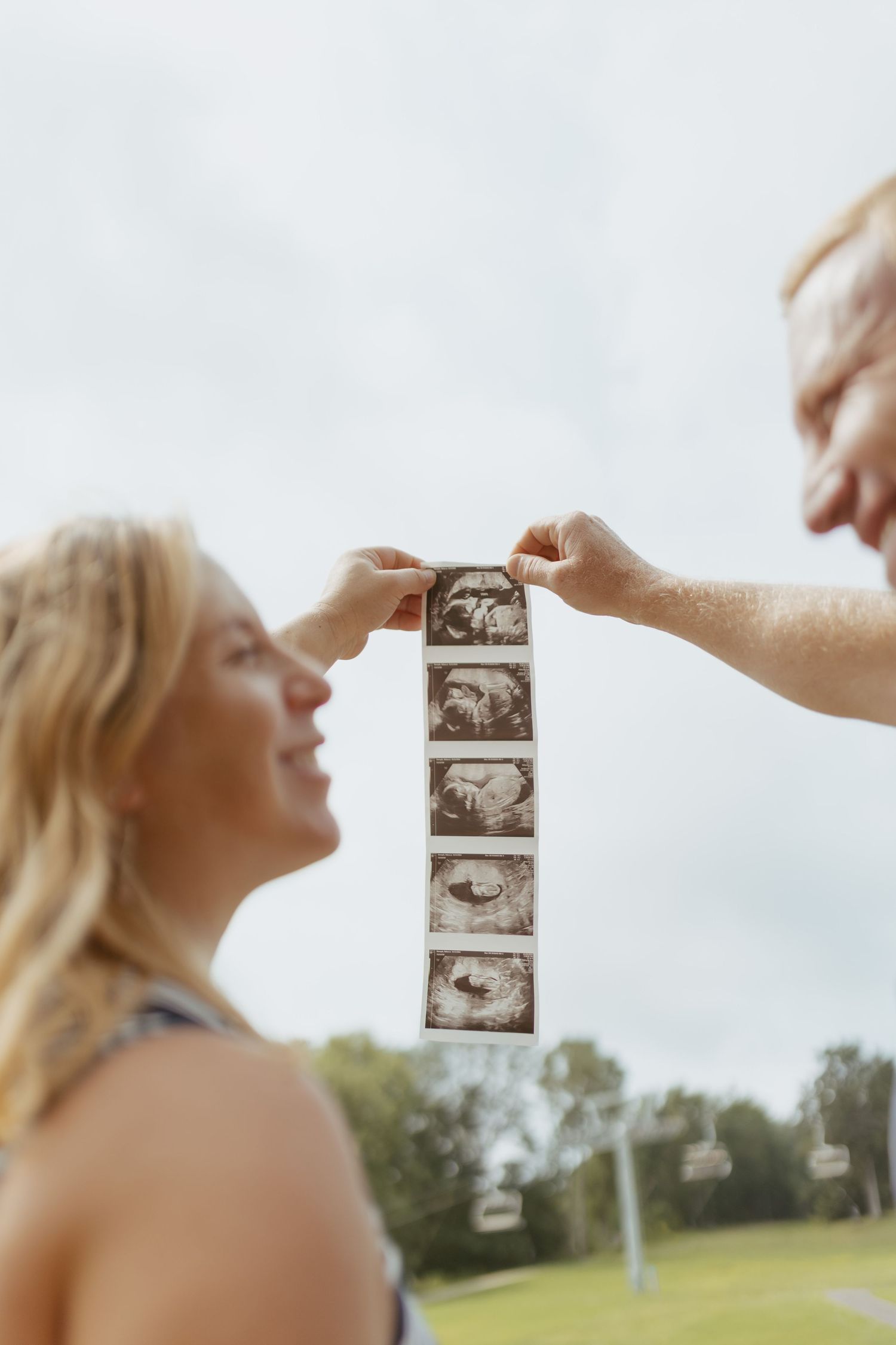 Couple holding ultrasound sonogram strip outdoors, smiling woman with blonde hair in pregnancy announcement photo.