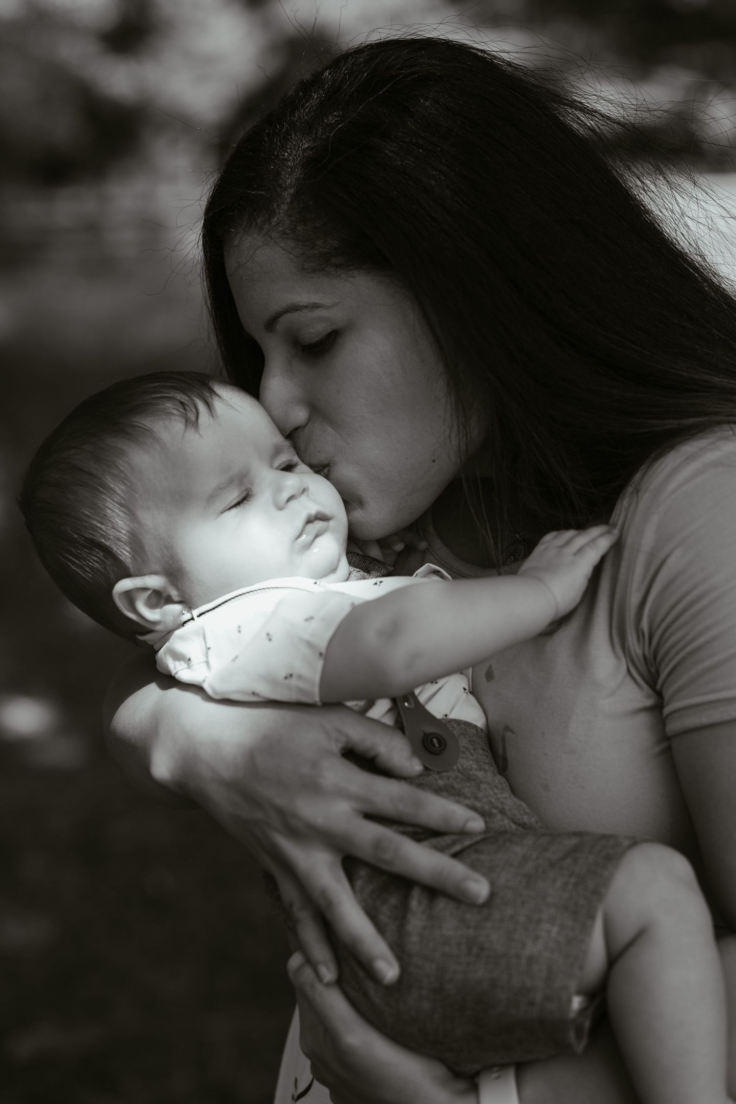 Black and white photo of a young mother tenderly kissing her sleeping newborn baby while cradling it in her arms.