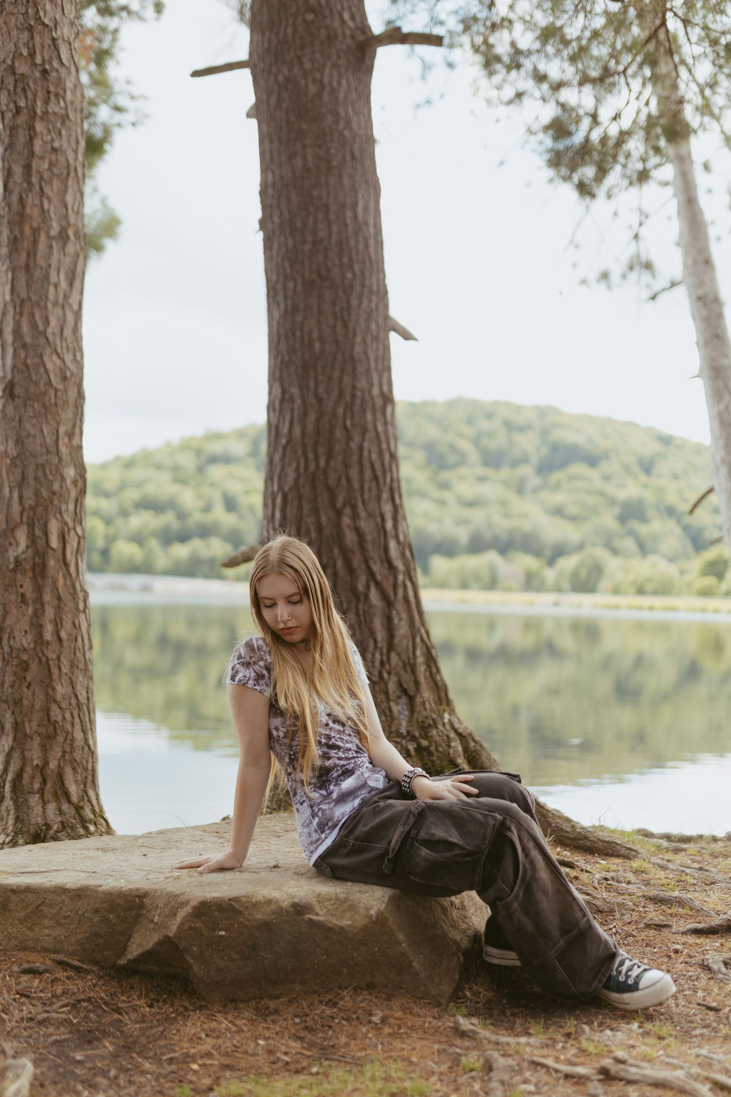 Young blonde woman sitting on a rock by a serene lake, surrounded by pine trees and lush green hills.