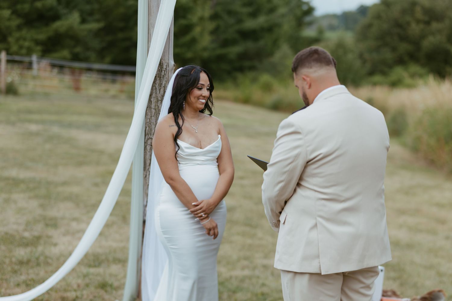 Bride in white gown smiling at groom in beige suit reading vows outdoors at rustic wedding ceremony.