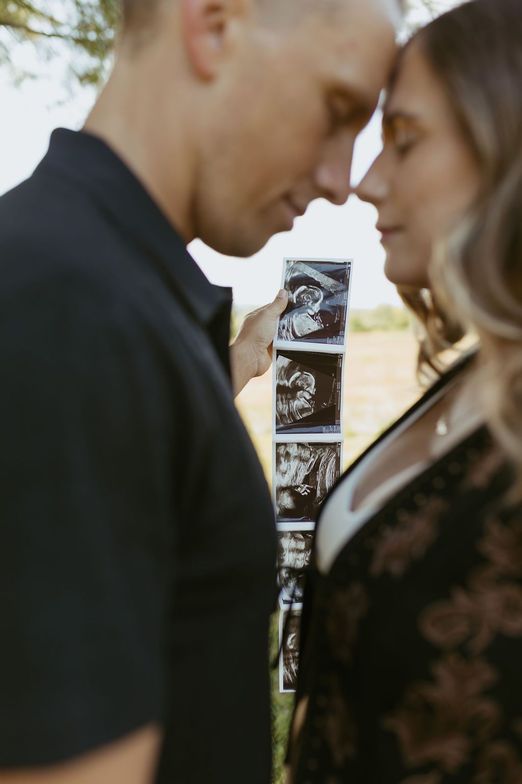 Couple touching foreheads while holding ultrasound photos outdoors, announcing pregnancy in a tender moment.