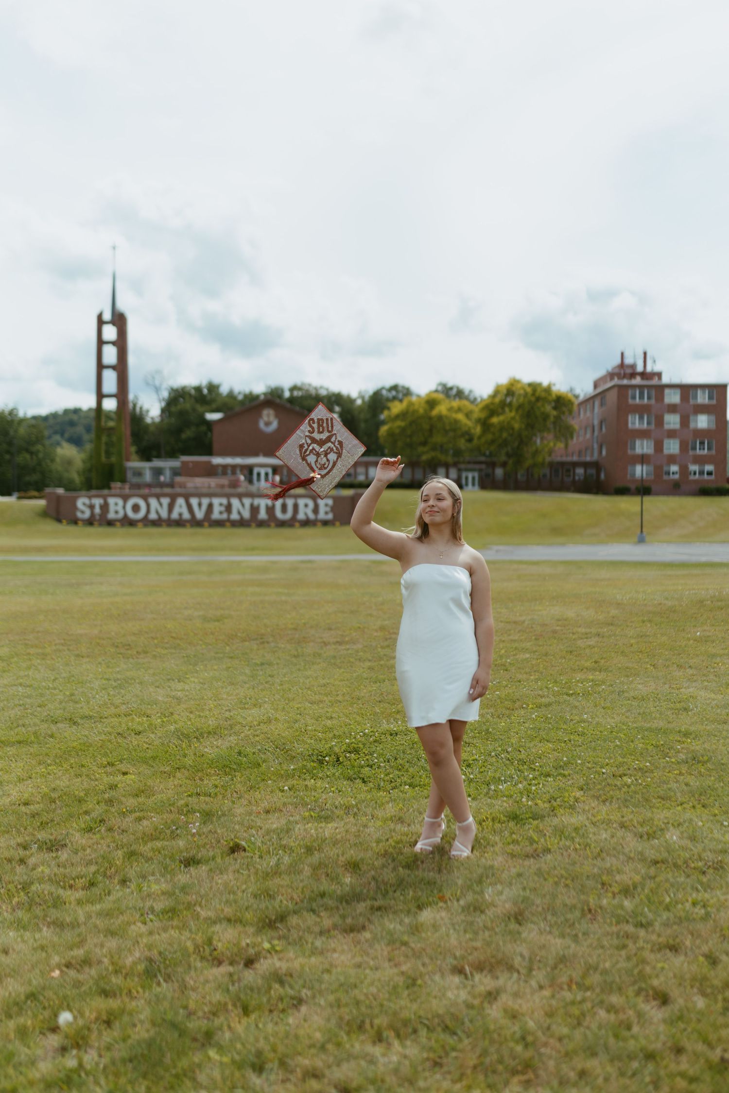 Young woman in white dress posing on green lawn at St. Bonaventure University campus on cloudy day.