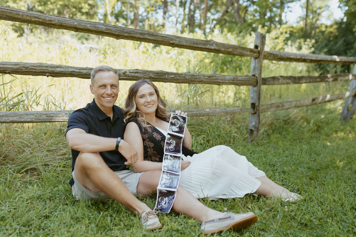 Happy couple sitting on grass by wooden fence, holding ultrasound photos, announcing pregnancy outdoors.