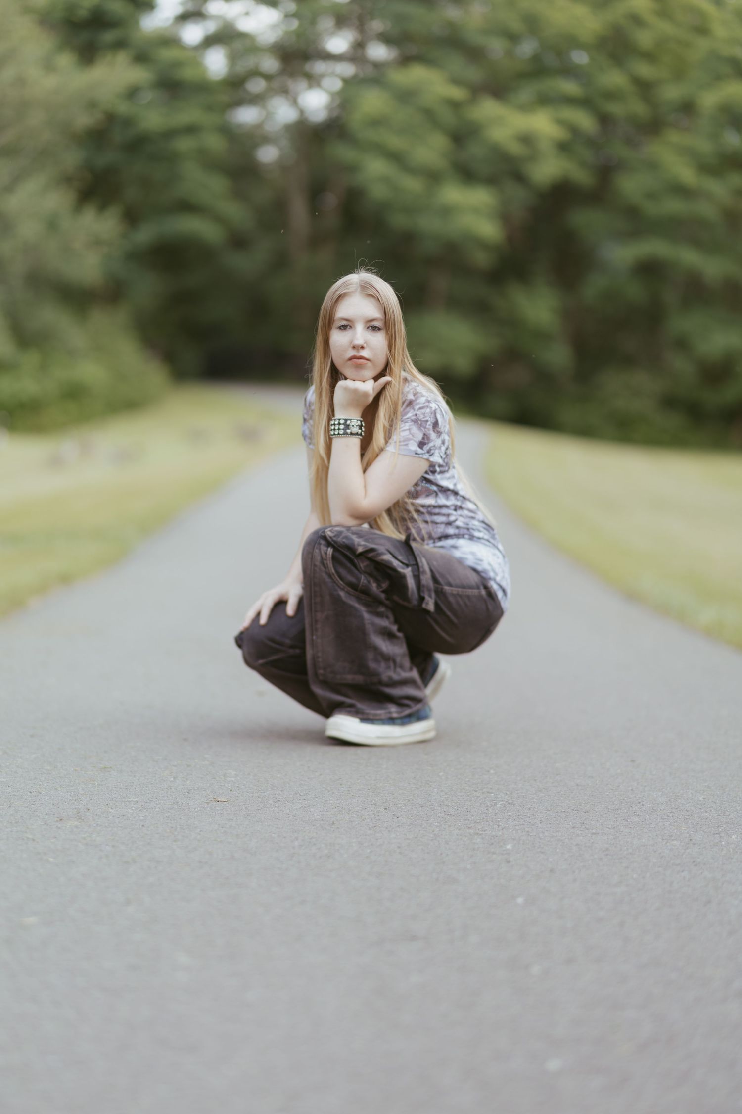 Young woman crouching on a tree-lined path, wearing gray pants and a floral top, resting her chin on her hand.