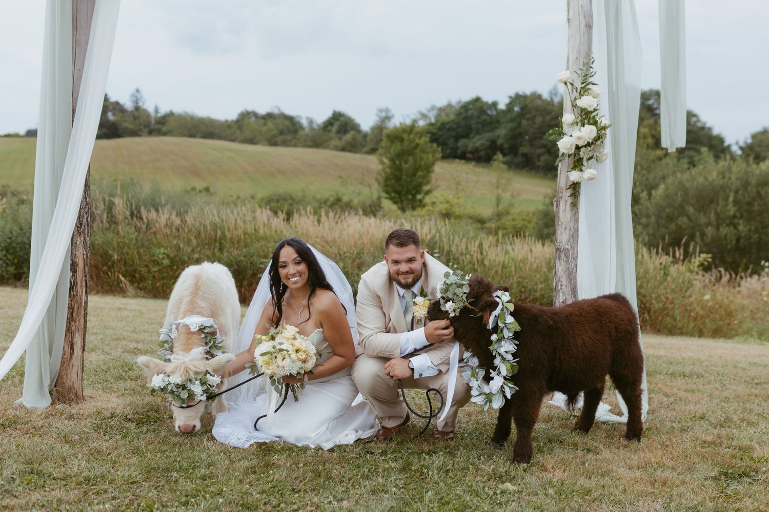 Bride and groom kneeling beside a flower-adorned brown calf at an outdoor rustic wedding ceremony.