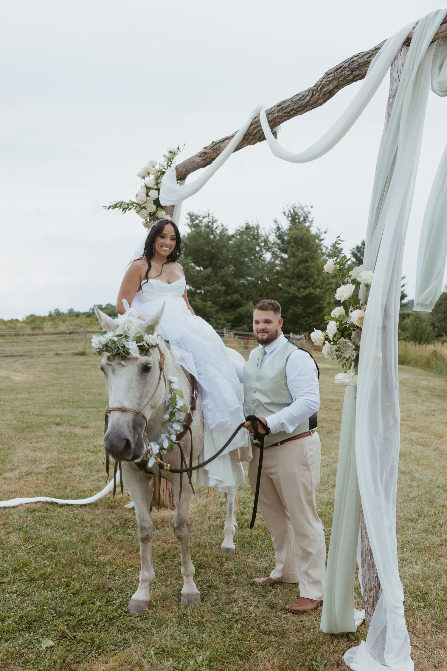 Bride sits on a white horse adorned with floral crown as groom leads her past a rustic wedding arch outdoors.