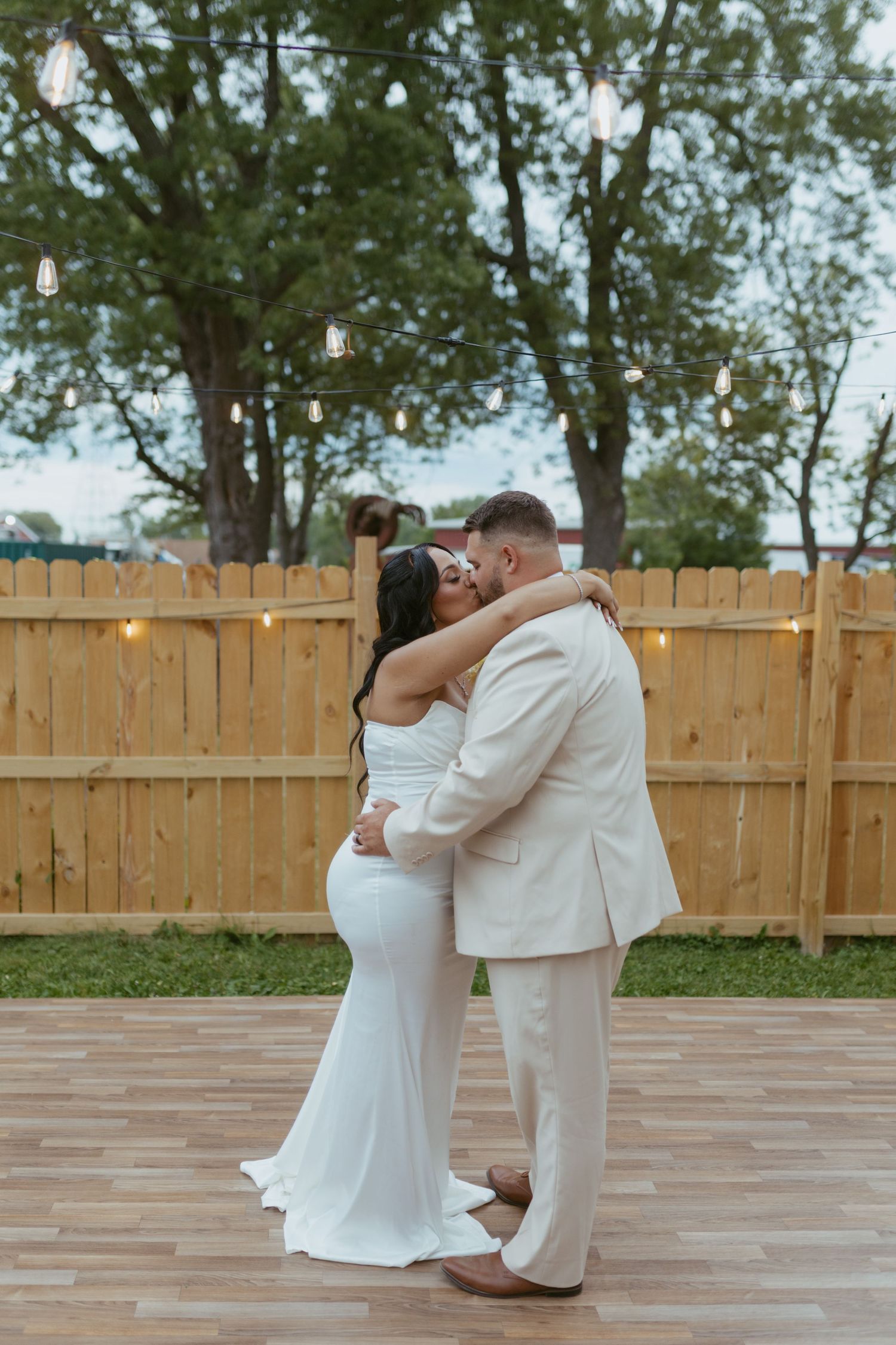 Bride and groom sharing a kiss during their first dance outdoors, surrounded by string lights and a wooden fence backdrop.