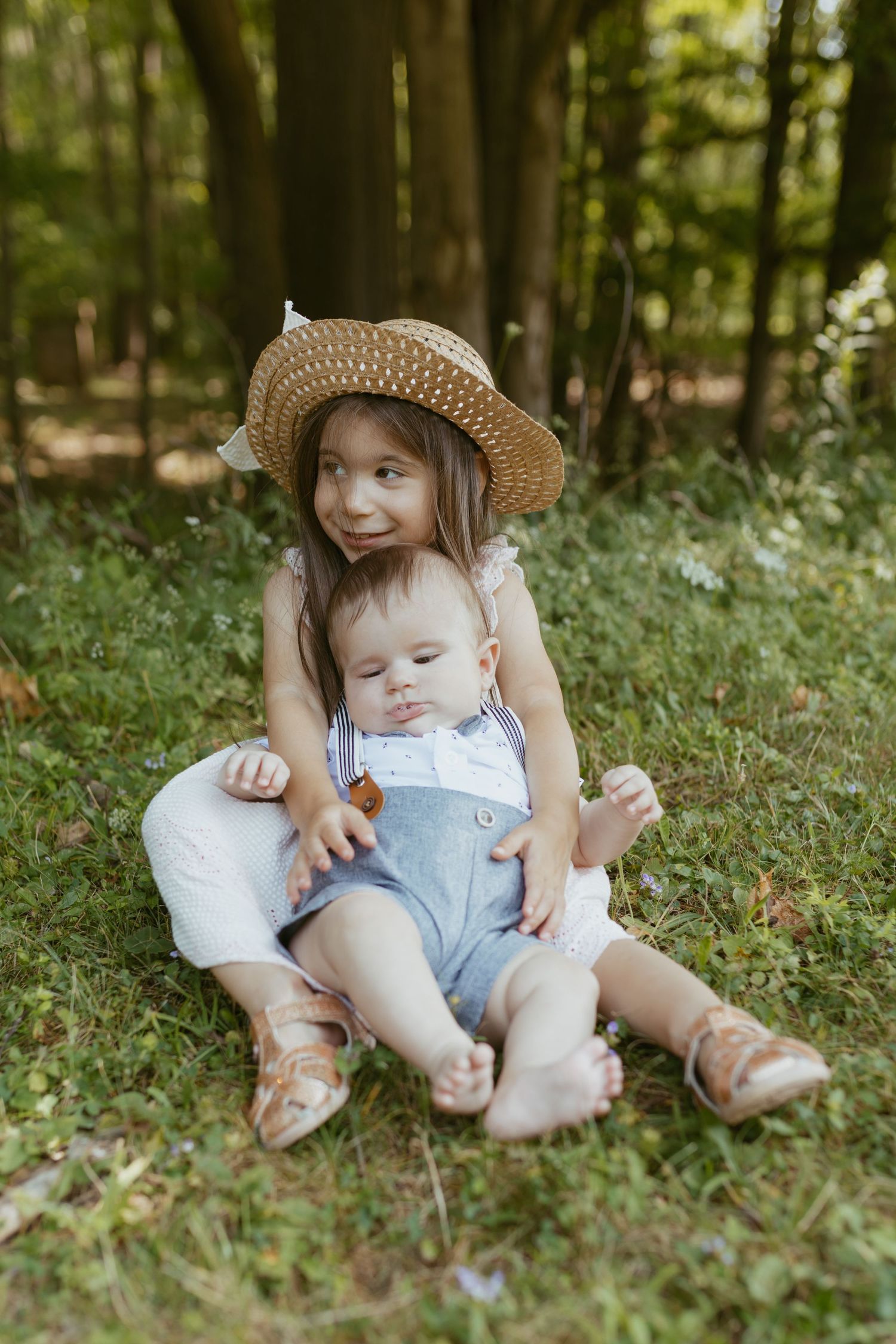A young girl in a straw hat sits on grass in a forest, lovingly holding a baby in blue overalls.