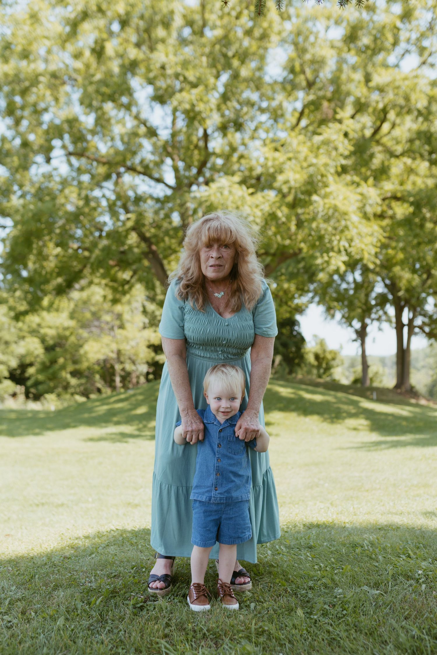 Woman in teal dress holds toddler in denim romper outdoors on green grass with trees in background.