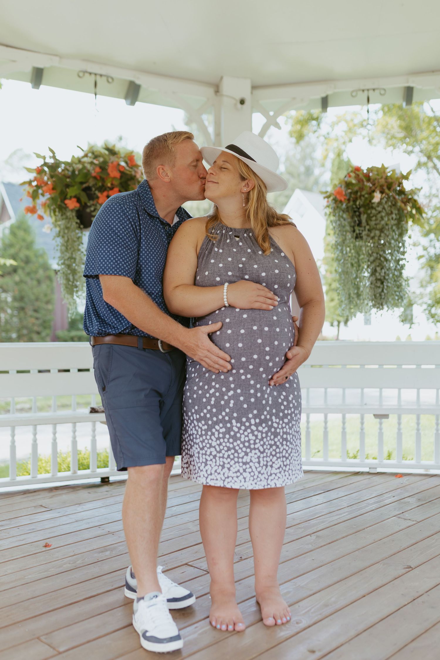 A couple shares a kiss in a gazebo, the woman in a polka dot dress cradling her pregnant belly, surrounded by hanging flowers.