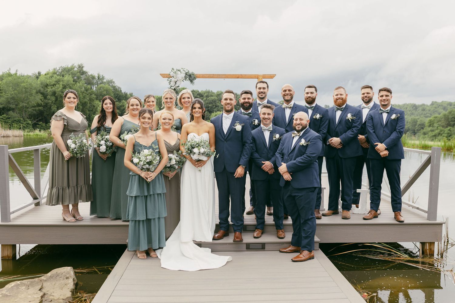 Wedding party members in navy suits and sage green dresses pose together on a scenic overlook.