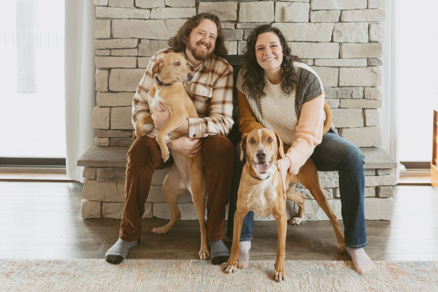 A smiling couple sits with two golden-brown dogs in front of a stone fireplace in a cozy home setting.