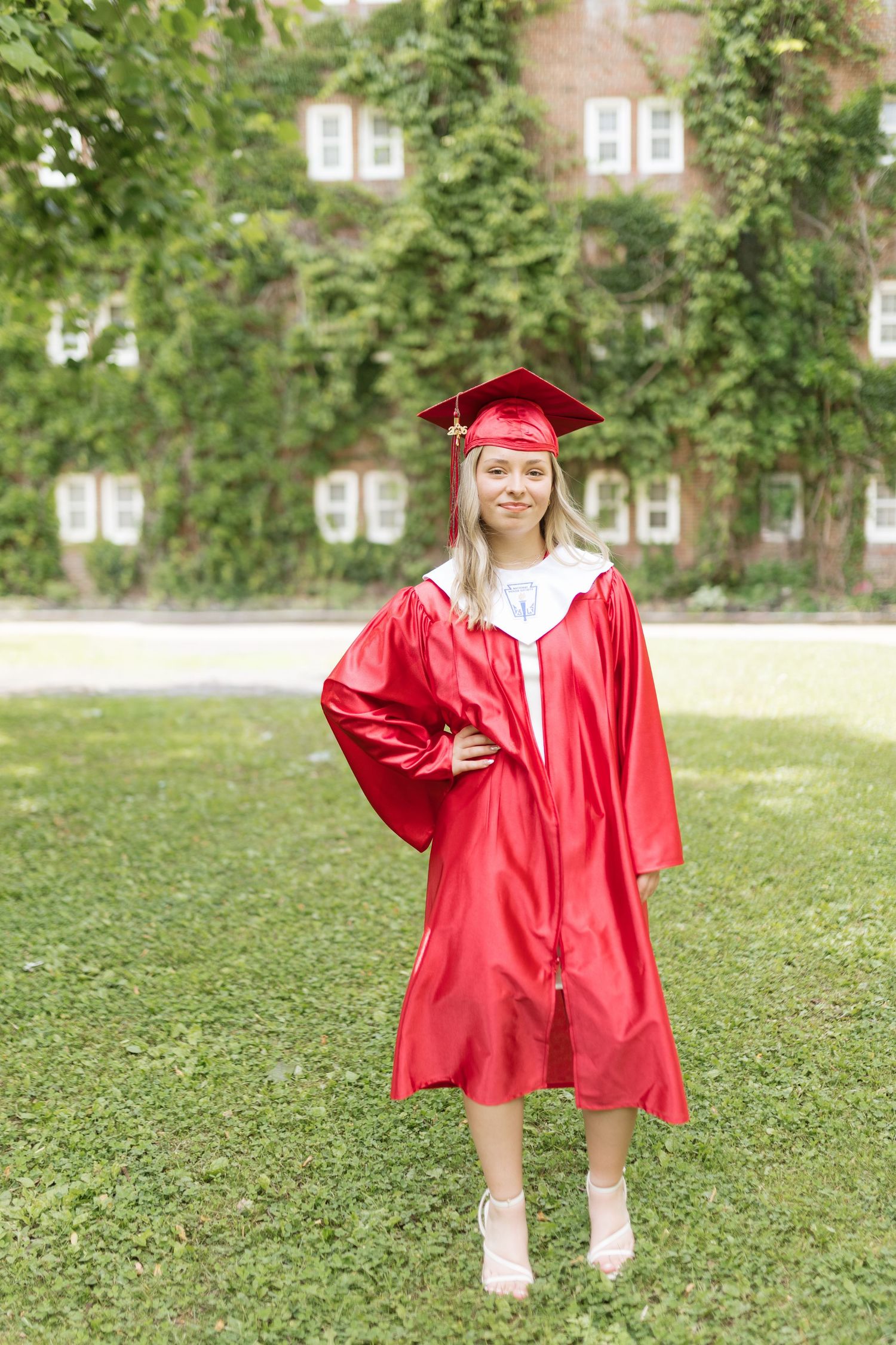 Young woman in red graduation cap and gown smiling outdoors on green lawn with ivy-covered brick building behind her.