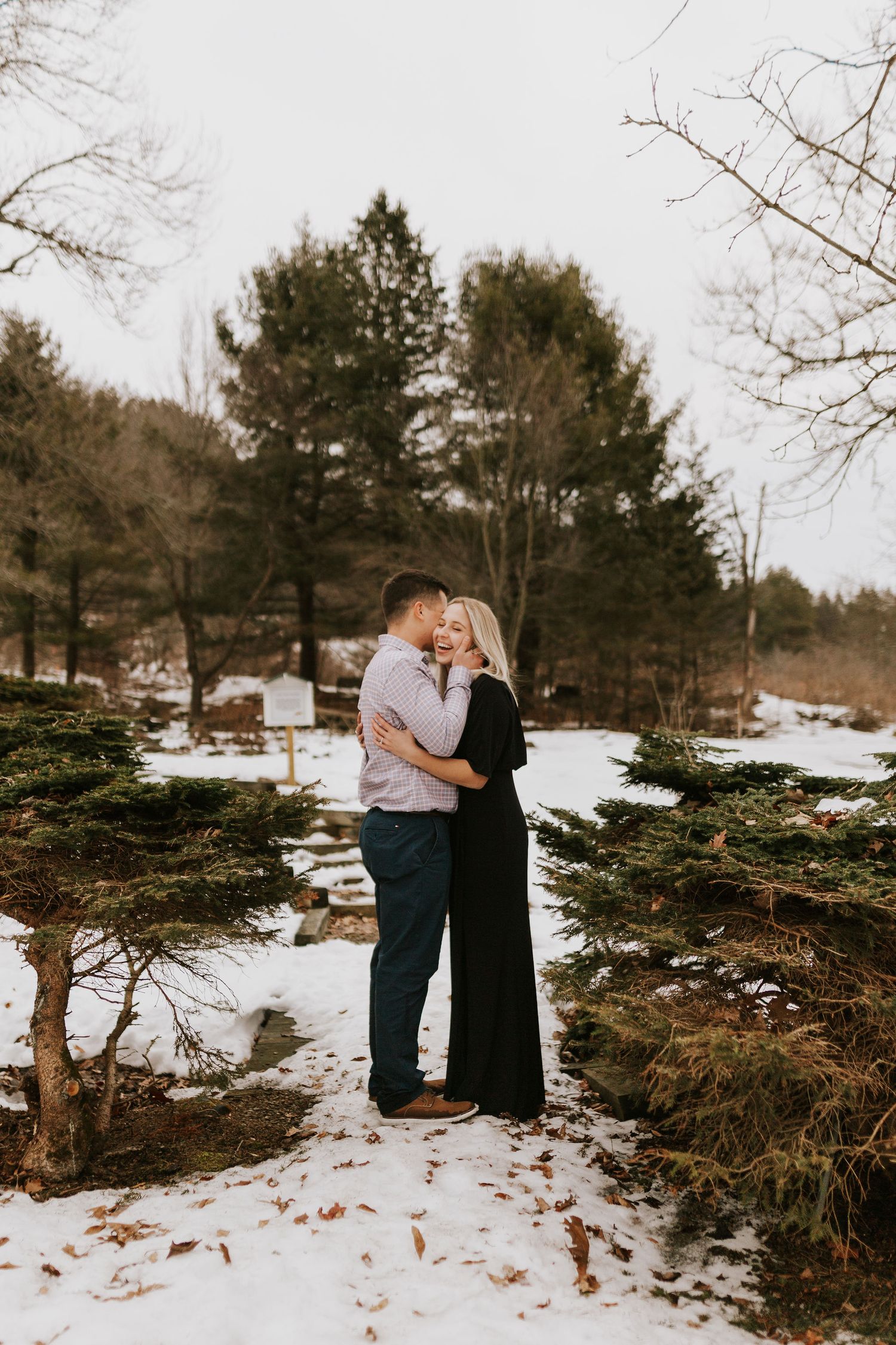 Couple embracing and almost kissing in a snowy winter forest setting with evergreen trees and bare branches.