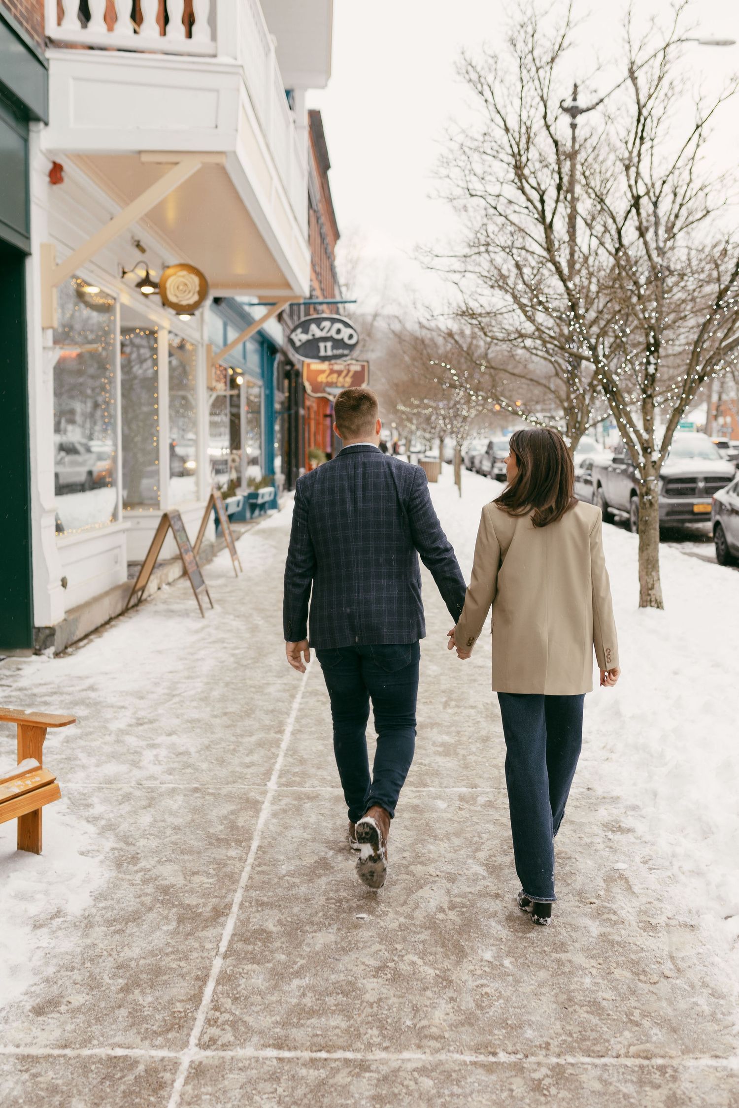 Couple holding hands walking on snowy sidewalk past shops in winter, man in plaid blazer and woman in beige coat.