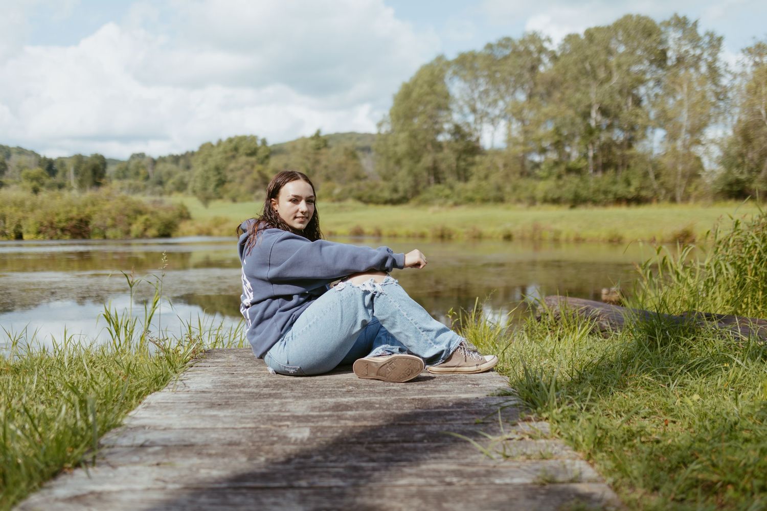 Young woman in blue hoodie and jeans sitting on wooden dock by a serene lake surrounded by lush green trees.
