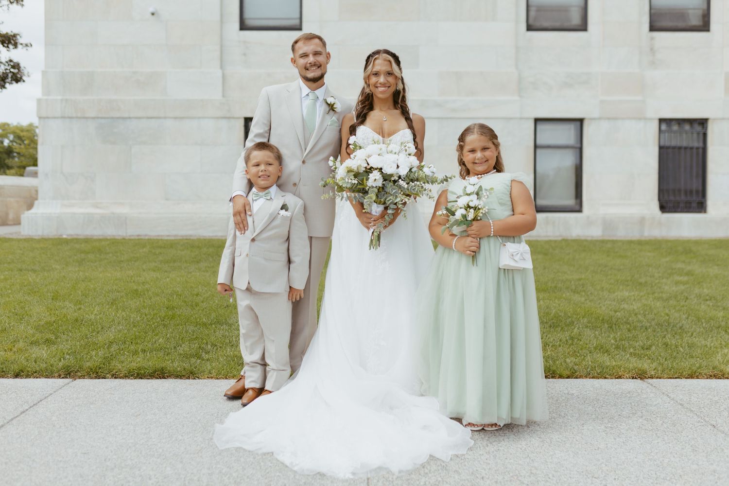 Wedding couple in beige and white attire pose with flower girl in mint dress and ring bearer outside a building.
