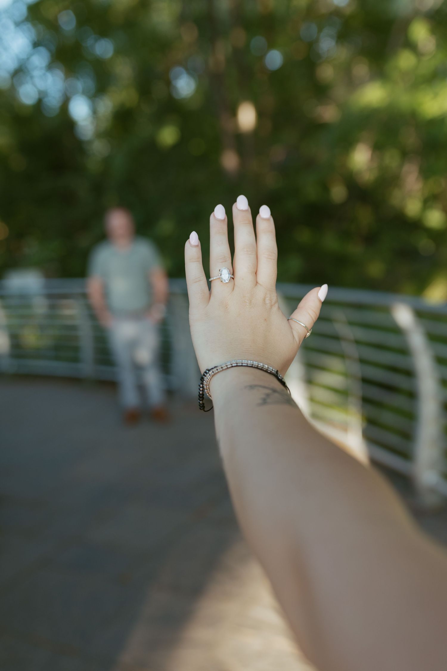 Close-up of engagement ring on womans hand with man standing on bridge background.