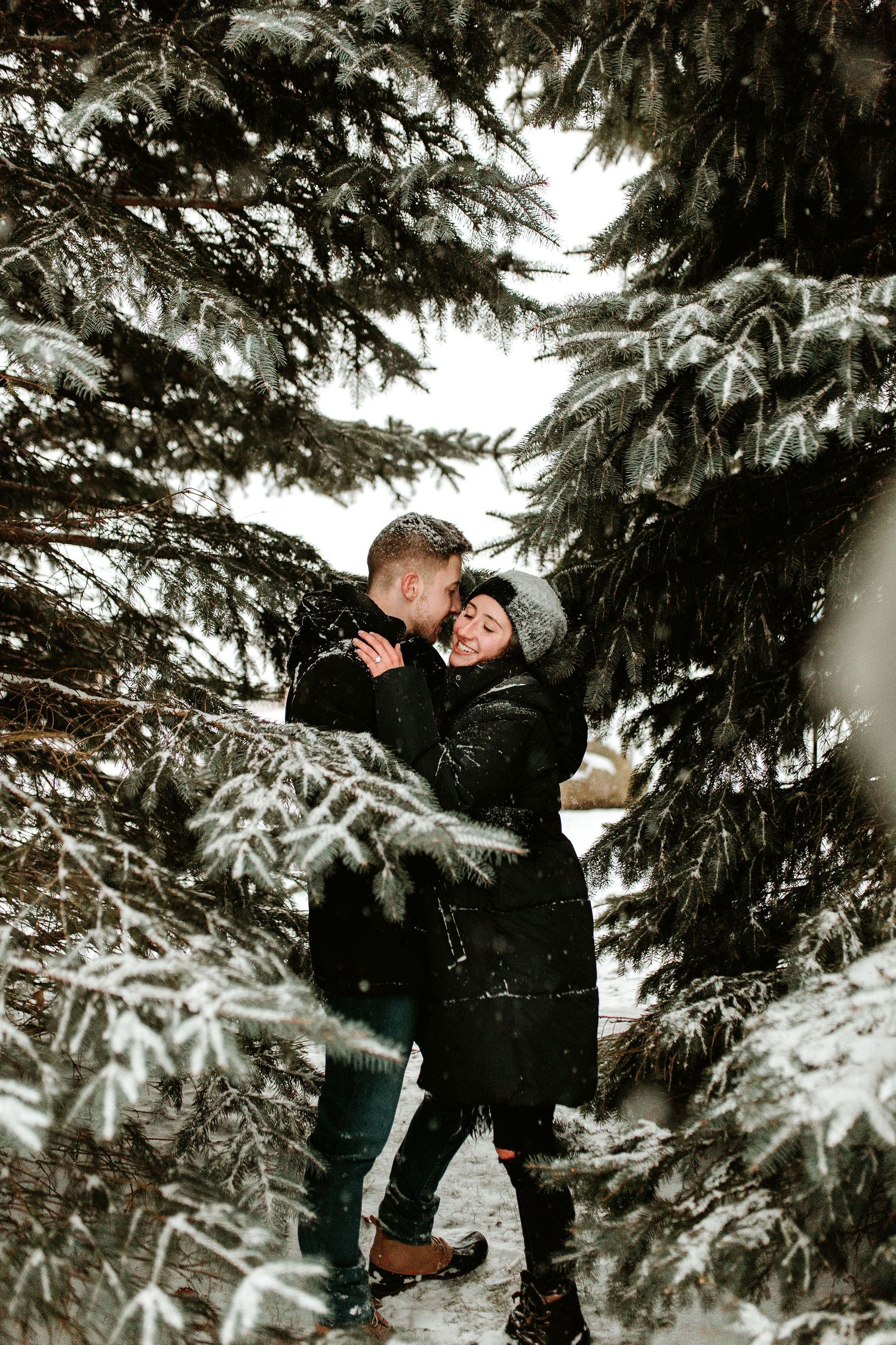 Couple embracing and kissing among snow-covered evergreen trees in a winter forest, wearing black coats.