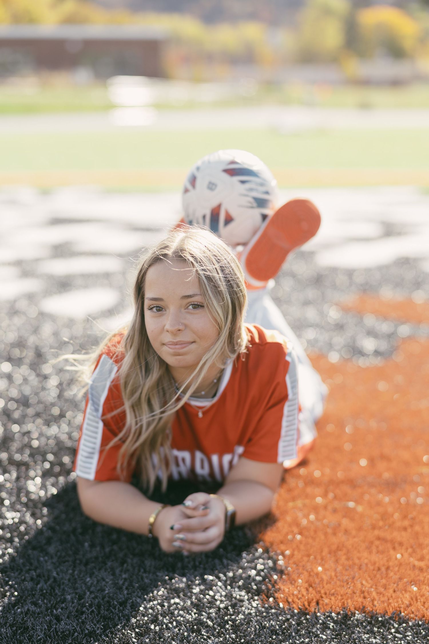 Young woman in red Warriors jersey lying on turf field with soccer ball, senior portrait photo.