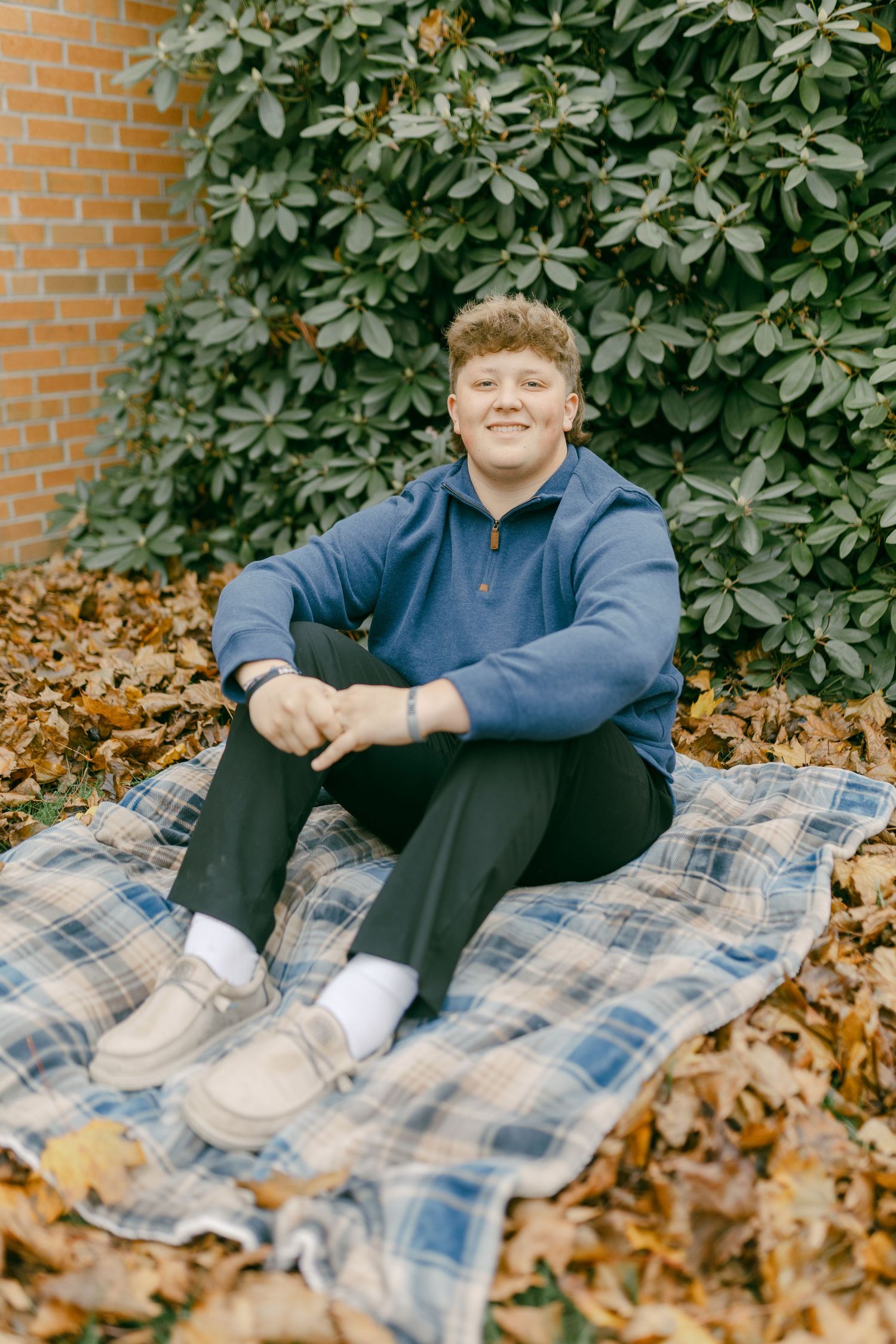Smiling young man in blue pullover sitting on plaid blanket among fall leaves near brick wall.