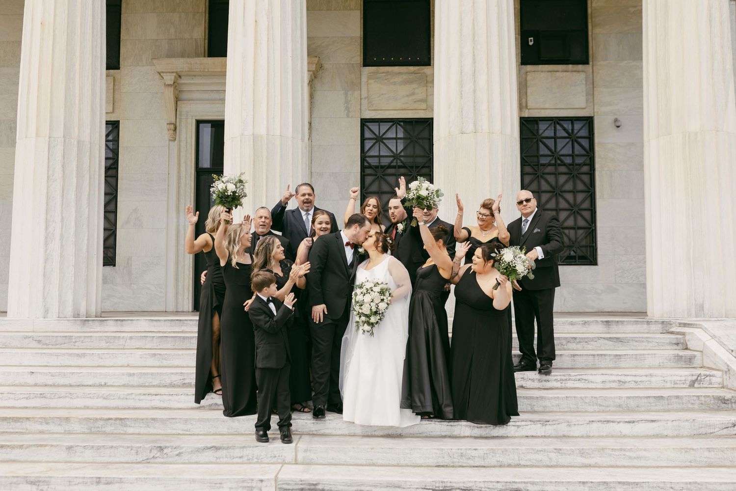 Wedding party in black attire celebrating on steps of a grand columned building with bride in white gown.