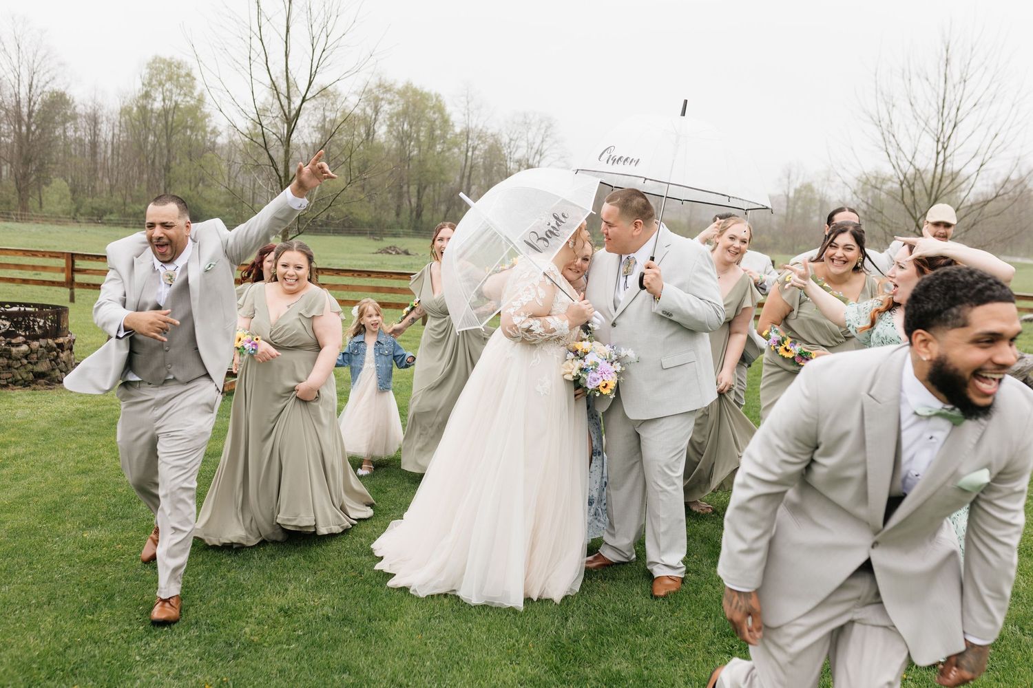 Wedding party celebrates outdoors in the rain, bride and groom share umbrella while bridal party laughs and walks across green lawn.