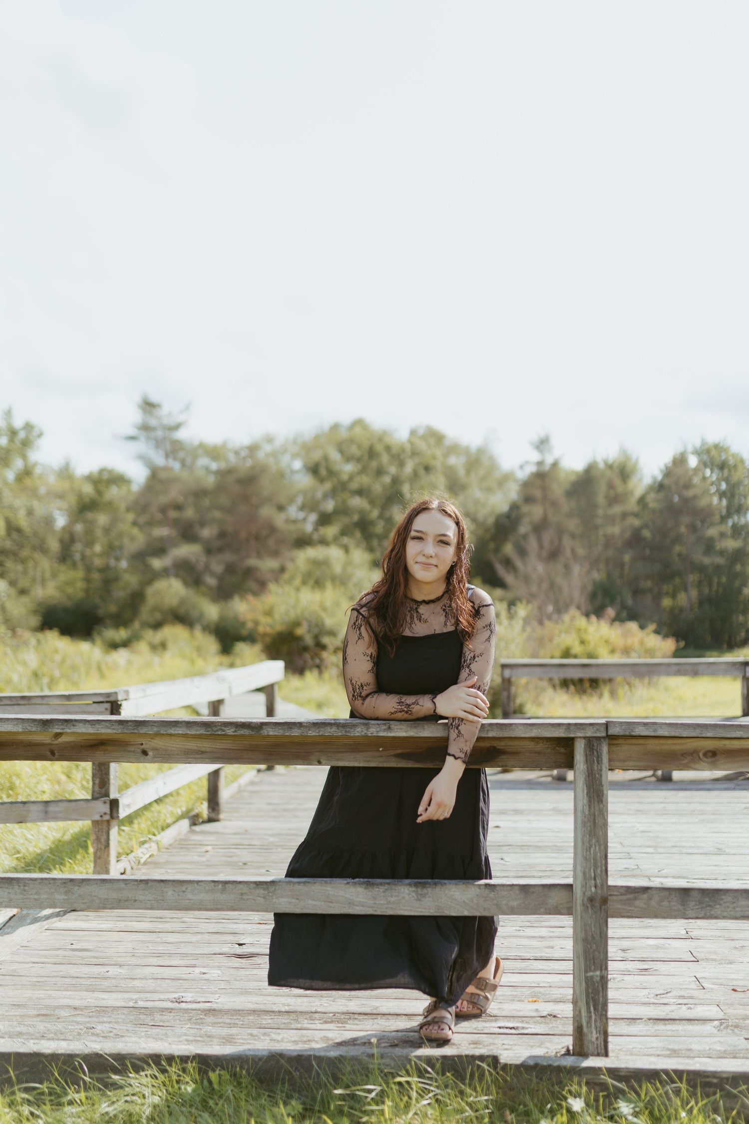 Young woman in black dress leaning on wooden boardwalk railing surrounded by lush greenery on a cloudy day.