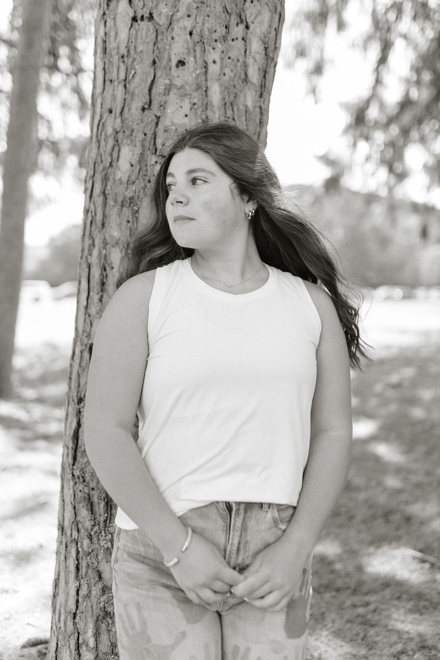 Black and white photo of a young woman leaning against a tree in a park, wearing a white tank top.