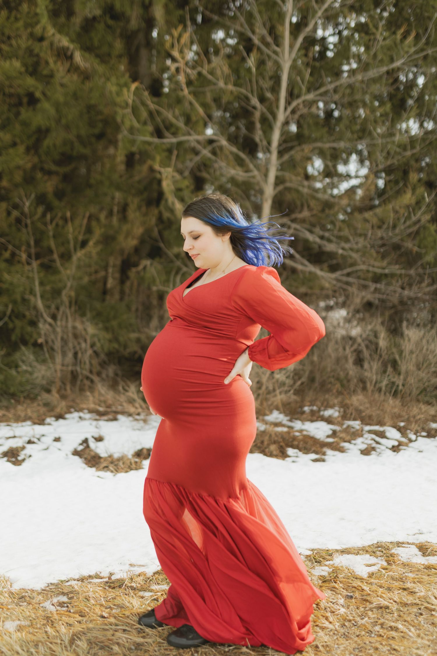 Pregnant woman in red gown with blue hair poses outdoors in snowy winter setting among evergreen trees.