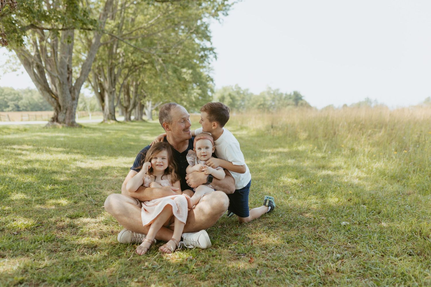 Family of five sitting on grass in a park, parents hugging children outdoors on a sunny day.