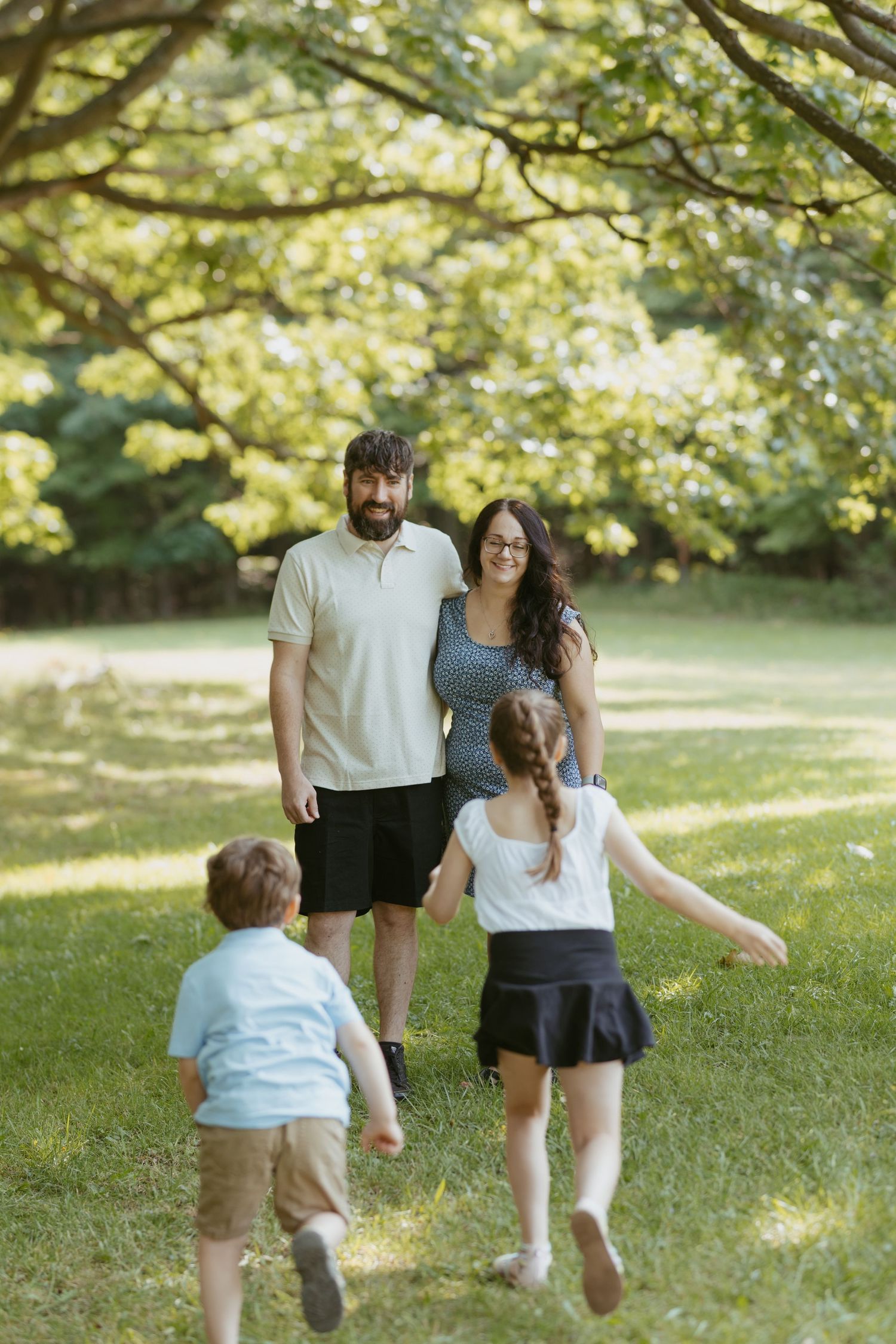 A happy family of four enjoying a sunny day in a lush green park, with two children running toward their smiling parents.
