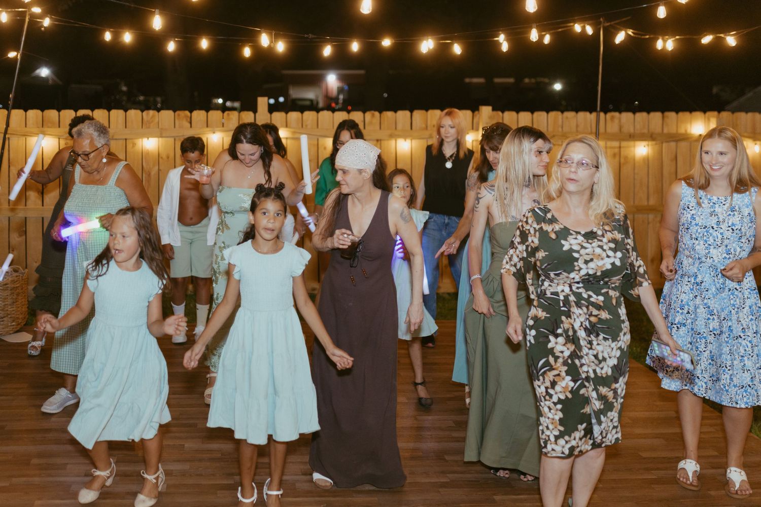 Guests dancing at an outdoor wedding reception under string lights on a wooden dance floor.