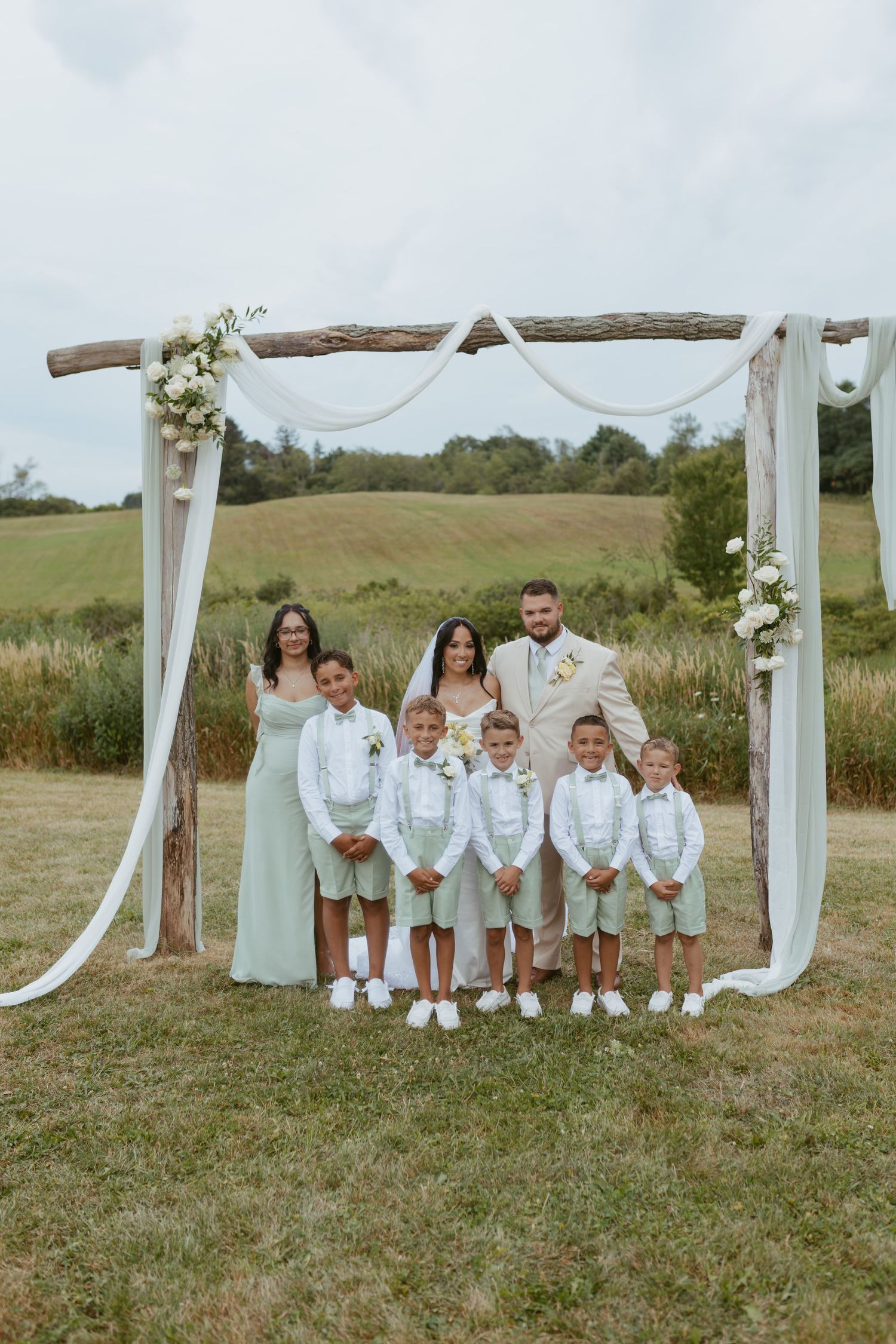 Wedding couple with children in sage green outfits posing under rustic wooden arch with white draping outdoors.