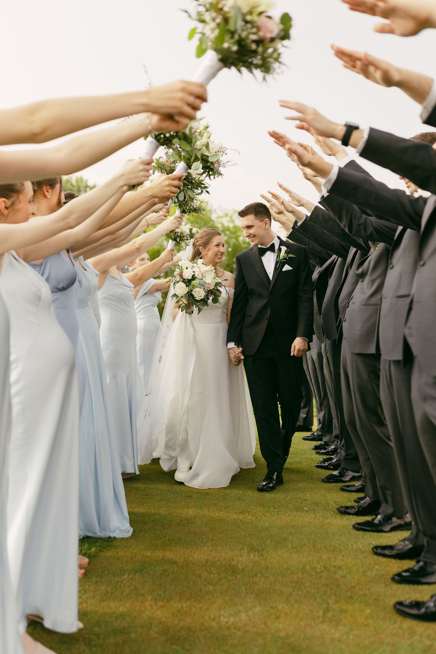 Wedding party forms a tunnel with raised arms as couple walks through on grassy lawn.