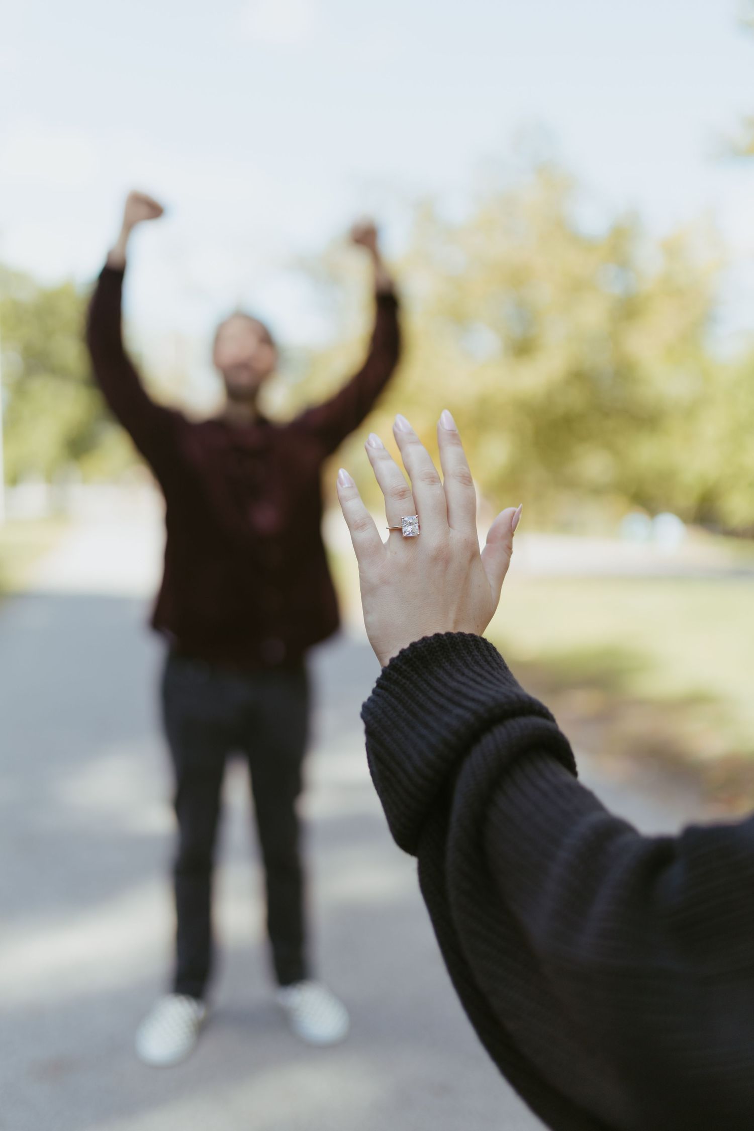Woman showing off engagement ring with excited fiancé celebrating in background at outdoor park.