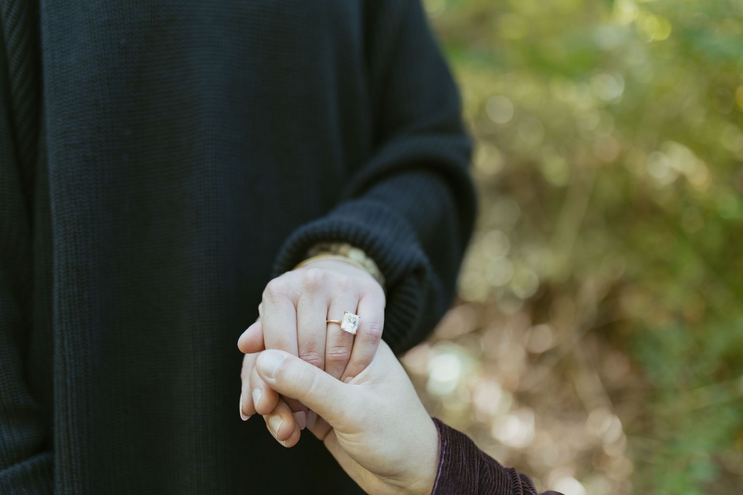 Two people holding hands outdoors, one wearing a gold engagement ring, with a blurred green nature background.
