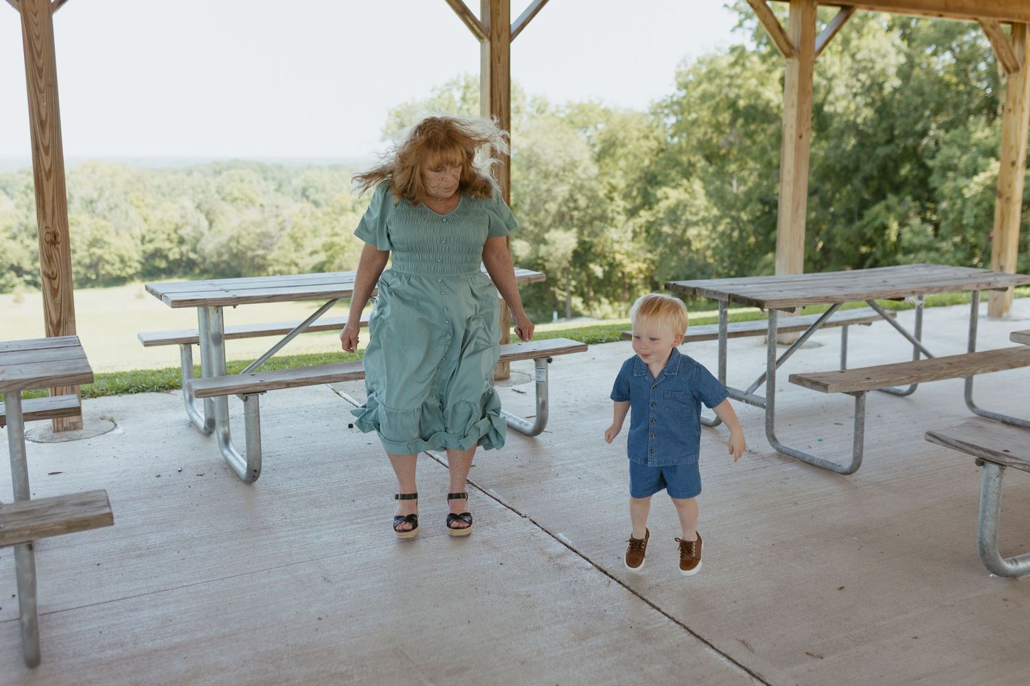 A woman in a green dress and a toddler boy in blue romper playing under a park pavilion with picnic tables.