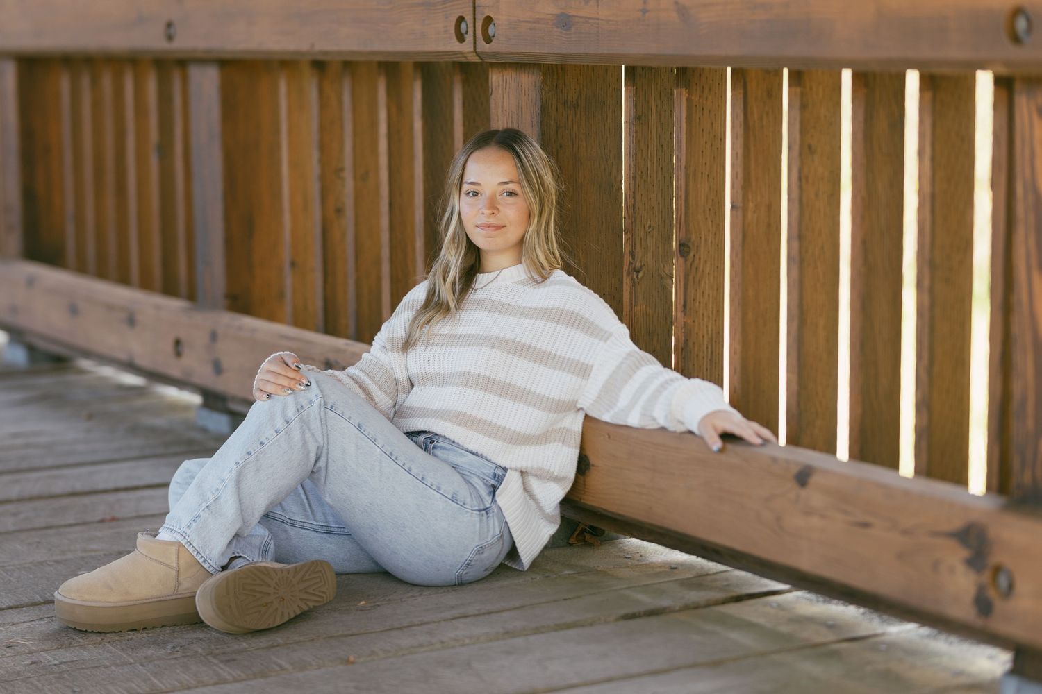 Young woman in striped sweater and jeans sitting on wooden bridge deck, smiling casually for senior portrait.