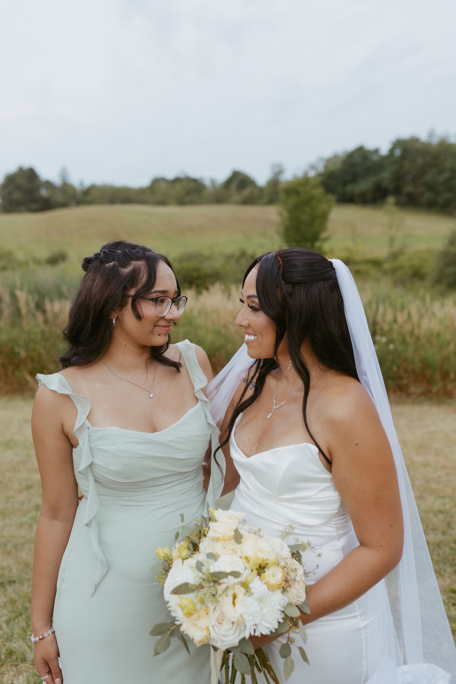 A bride in a white gown and veil holds a bouquet while smiling at her bridesmaid in a sage green dress outdoors.