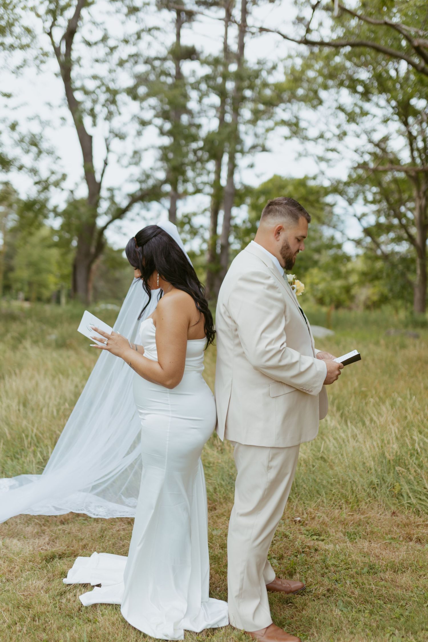 Bride and groom stand back-to-back reading vows in a lush outdoor setting, she in white gown with veil, he in beige suit.